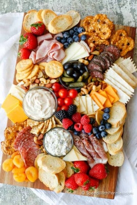 cheeses , meats , breads, nuts , fruits and pickles with olives on a board to show How to Make a Charcuterie Board