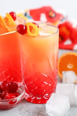 two glasses of Tequila Sunrise with cherries , oranges and ice cubes