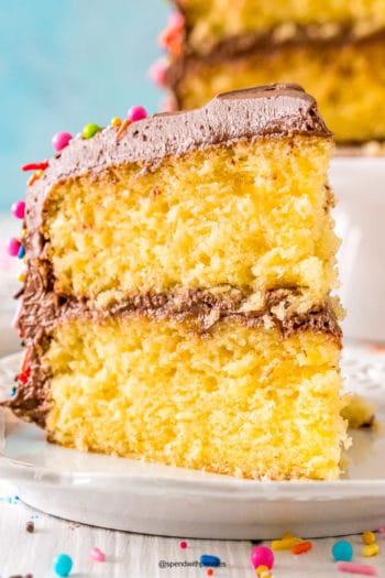 Close up photo of a slice of yellow cake with chocolate frosting on a white plate.